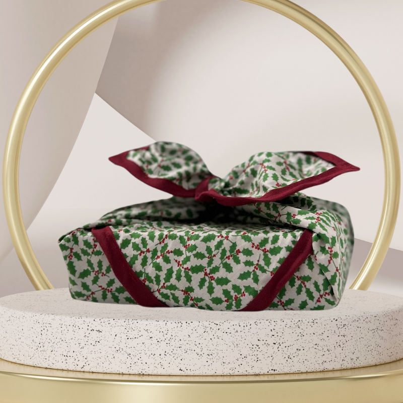 A luxury reusable fabric gift wrap with a holly pattern, featuring red berries and green leaves on a white background, eco friendly, personalised through embroidery 